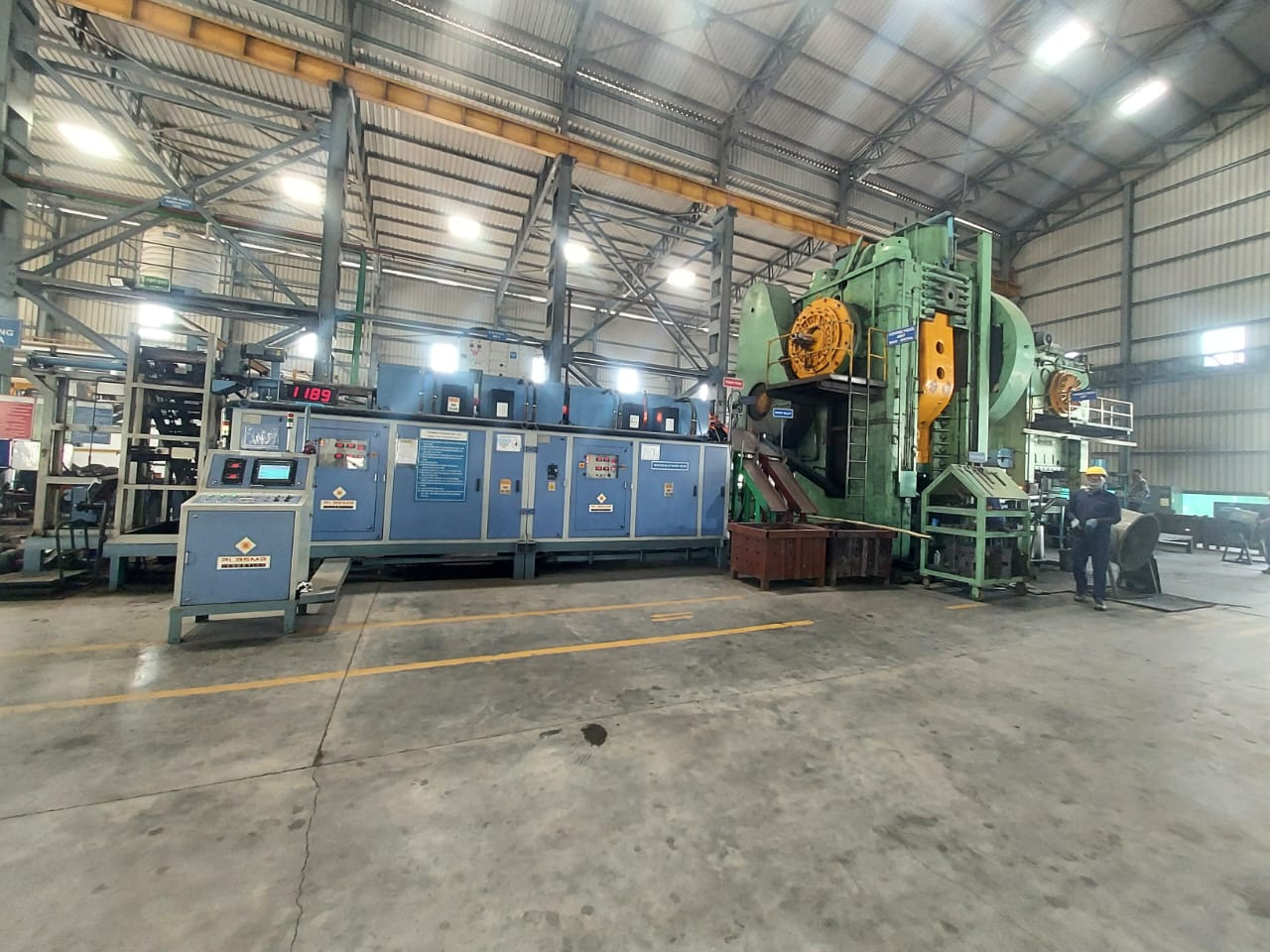 Forging press bay at our Pune partner facility, India · induction heater, blue control panels, operator in PPE during a closed die run